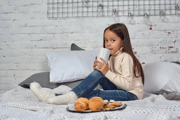 Menina bonita 6 anos de idade em uma camisola branca e jeans. Criança no quarto com uma cama, comer croissant e beber chá ou cacau. — Fotografia de Stock