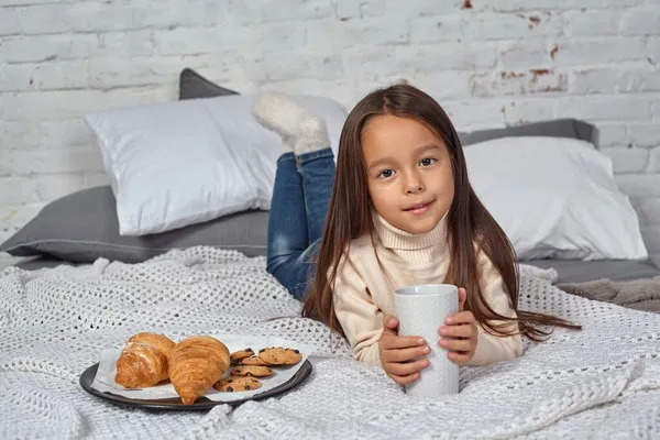 Jolie petite fille de 6 ans en pull blanc et jean. Enfant dans la chambre avec un lit, manger croissant et boire du thé ou du cacao. — Photo