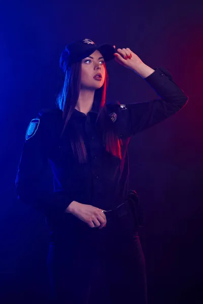 Une policière sérieuse pose pour la caméra sur un fond noir avec un rétroéclairage rouge et bleu . — Photo