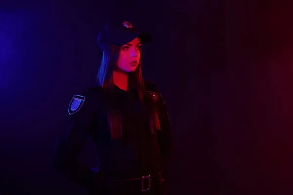 Allvarlig kvinnlig polis är poserar för kameran mot en svart bakgrund med röd och blå bakgrundsbelysning. — Stockfoto