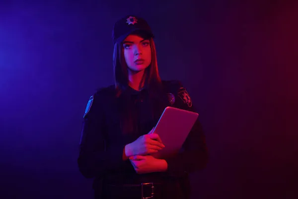 Sério policial feminino está posando para a câmera contra um fundo preto com backlighting vermelho e azul . — Fotografia de Stock