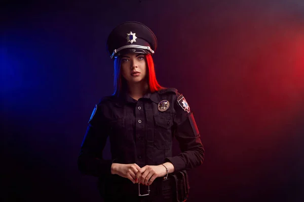 Sério policial feminino está posando para a câmera contra um fundo preto com backlighting vermelho e azul . — Fotografia de Stock