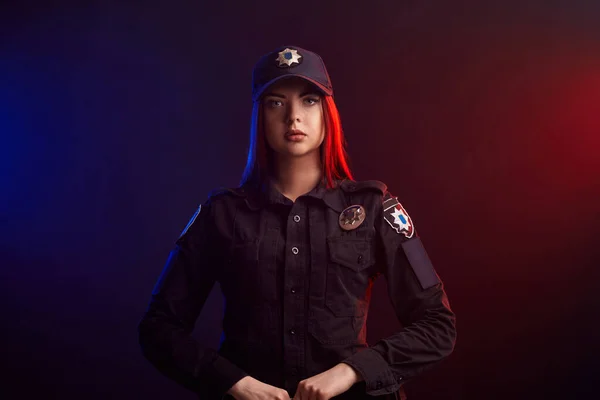Mujer policía seria posando para la cámara sobre un fondo negro con retroiluminación roja y azul . — Foto de Stock