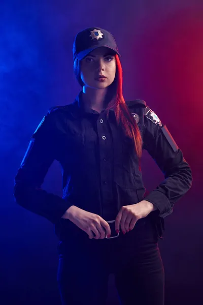 Mujer policía seria posando para la cámara sobre un fondo negro con retroiluminación roja y azul . —  Fotos de Stock