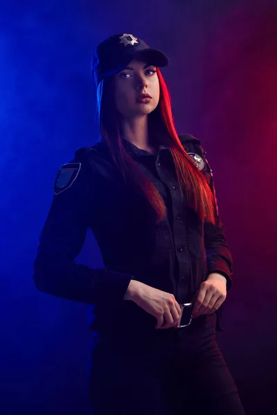 Sério policial feminino está posando para a câmera contra um fundo preto com backlighting vermelho e azul . — Fotografia de Stock