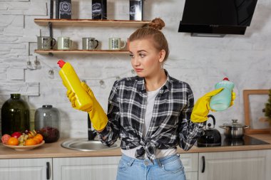 Deterjan ürünlerini karşılaştıran bir kadın elinde iki şişe tutuyor ve onları ev mutfağında kontrol ediyor..