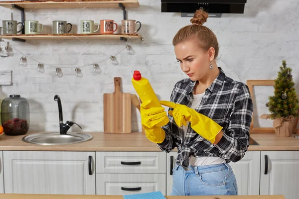 Επαγγελματική νεαρή γυναίκα φορώντας λαστιχένια προστατευτικά κίτρινα γάντια κρατώντας καθαριστικά μπουκαλιών στην κουζίνα. — Φωτογραφία Αρχείου