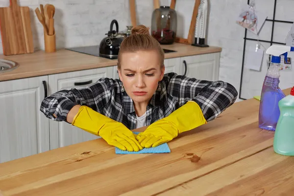 Νεαρή γυναίκα με προστατευτικά γάντια σκουπίζει ένα τραπέζι στην κουζίνα με ένα κουρέλι — Φωτογραφία Αρχείου