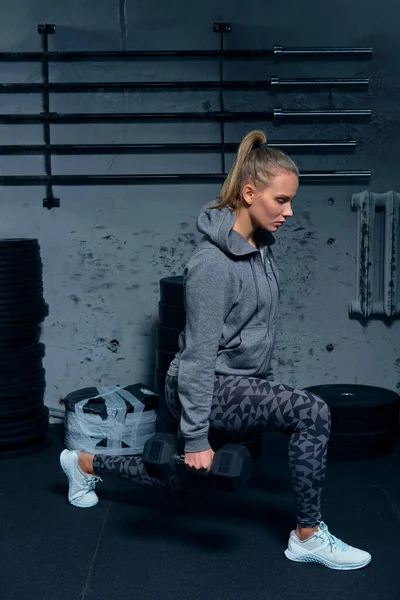Seitenansicht einer schönen jungen Fitnesstrainerin, die einen Sportanzug trägt und eine Übung mit Hanteln auf grauem Hintergrund macht — Stockfoto