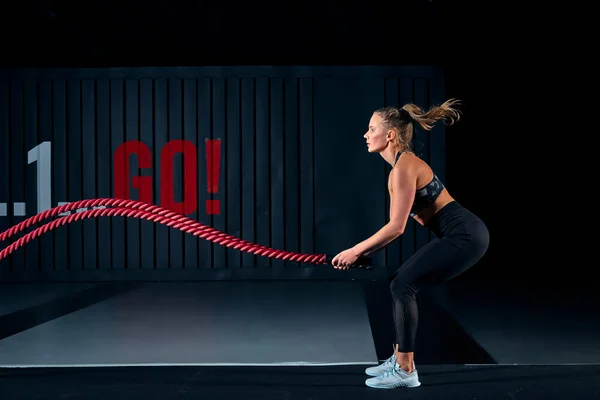 Mujer musculosa fuerte trabajando con cuerdas de batalla. Foto de mujer atractiva en ropa deportiva de moda en el gimnasio. Vista lateral. Fotos De Stock