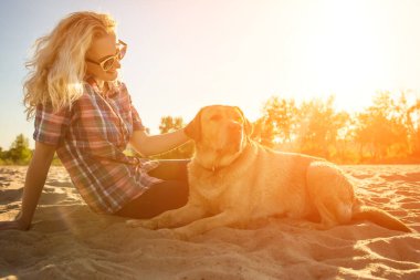 Golden Retriever köpeğiyle kumsalda oturan güneş gözlüklü güzel bir kadının portresi. Deniz kıyısında köpekli bir kız. Güneş parlaması