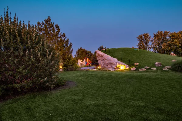 Twilight view of park with illuminated green lawn, decorative hill and children playground — Stock Photo, Image