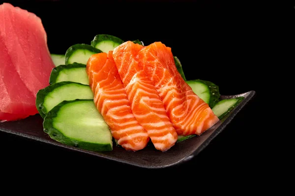 Closeup of raw salmon sashimi on platter with tuna and cucumber on black background — 图库照片