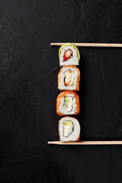 Τέσσερα ρολά σούσι με σολομό, χέλι, γαρίδες, αβοκάντο σε μαύρη επιφάνεια μεταξύ chopsticks — Φωτογραφία Αρχείου