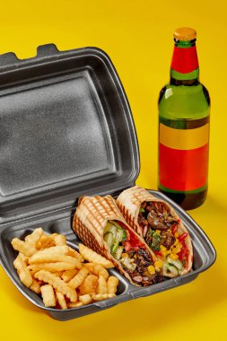 Takeaway container with vegan burrito and fries on yellow background with bottle of drink