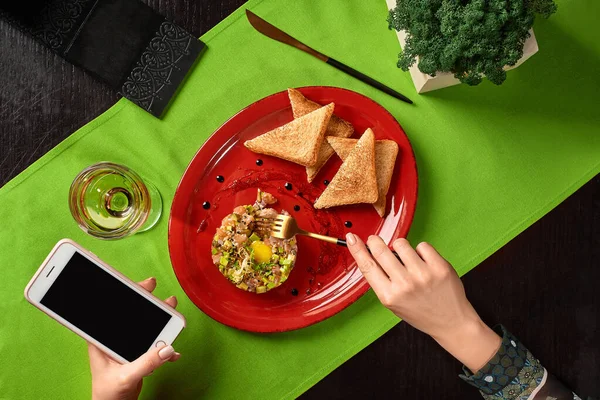 Woman hands holding phone and fork eating salmon tartare served with toasts — 스톡 사진