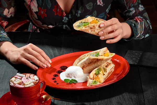 Mãos femininas segurando rolo de frutas em lavash fino servido com sorvete e xícara de cacau — Fotografia de Stock