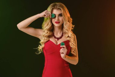 Kırmızı elbiseli ve kolyeli sarışın kadın. Gülümsüyor, yeşil çiplerle gözlerini kapamış, renkli arka planda poz veriyor. Poker, kumarhane. Yakın plan.