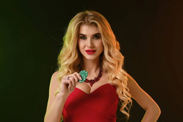 Ξανθό μοντέλο σε κόκκινο φόρεμα και κολιέ. Χαμογελάει, δείχνει δύο πράσινες μάρκες, ποζάροντας σε πολύχρωμο φόντο. Πόκερ, καζίνο. Κοντινό πλάνο — Φωτογραφία Αρχείου
