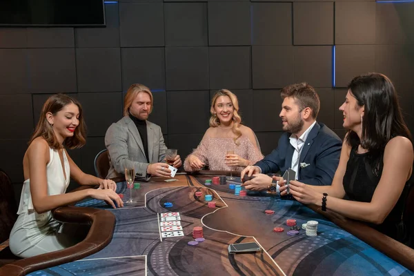 Mladí lidé hrají poker na přátelské párty, sedí u stolu s kartami a alkoholickými nápoji — Stock fotografie