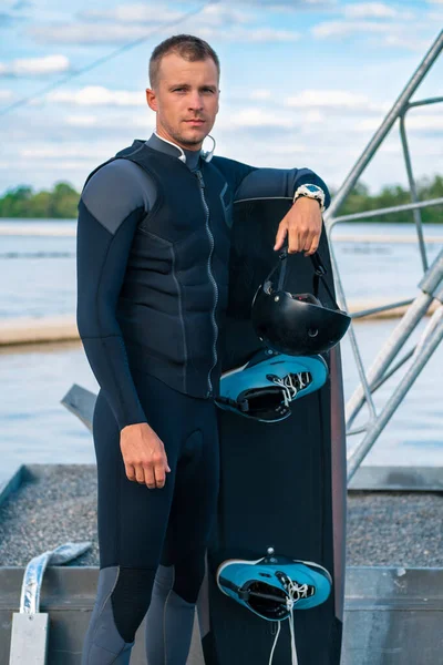 Selbstbewusster Wakeboarder im Neoprenanzug steht auf Pier an Bord gelehnt — Stockfoto