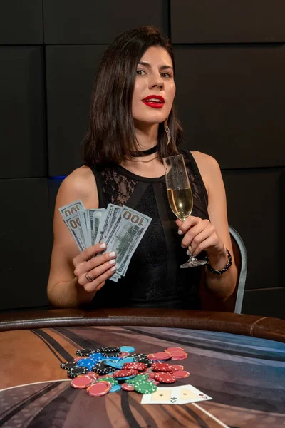 Šťastná mladá brunetka se sklenkou šampaňského a bankovkami sedící u pokerového stolu — Stock fotografie