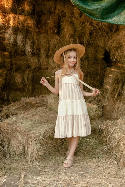 Романтическая девочка в светлом сарафане и соломенной шляпе, стоящей на сеновале — стоковое фото
