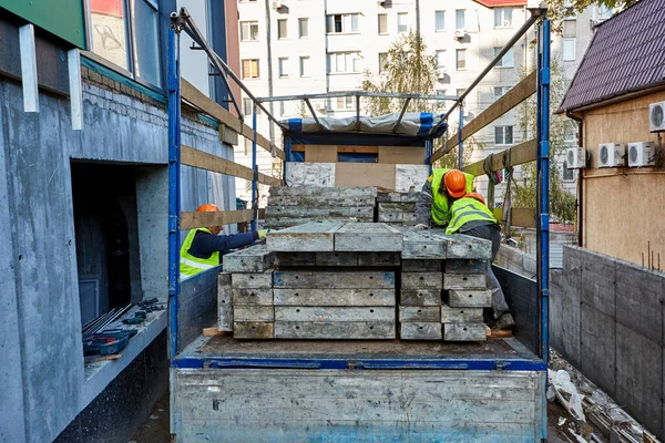 Професійні будівельники на робочому одязі та помаранчевих хардхатах розвантажують вантажівку матеріалами біля будівлі, що будується. — стокове фото