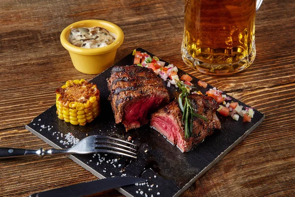 Gegrilltes Steak mit Mais mit Pilzsoße auf Schneidebrett und Bierkrug auf Holztisch — Stockfoto