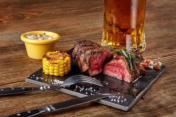 Grilled steak with corn with mushrooms sauce on cutting board and mug of beer on wooden table — Stock Photo, Image