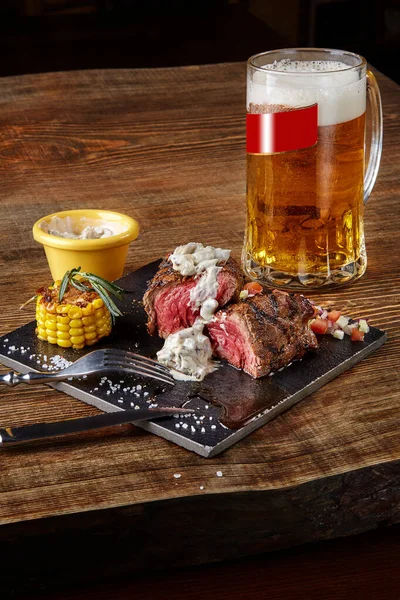Grilovaný steak s kukuřicí s houbovou omáčkou na řezací desce a džbánem piva na dřevěném stole — Stock fotografie