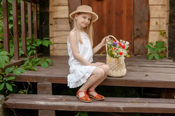 Speels tussen een meisje zitten op de drempel van het landhuis met mand van wilde bloemen — Stockfoto