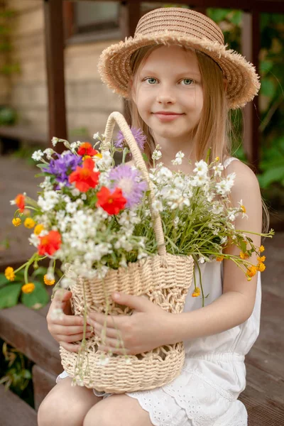 Усміхнена дівчинка з кошиком польових квітів сидить на порозі сільського будинку. — стокове фото