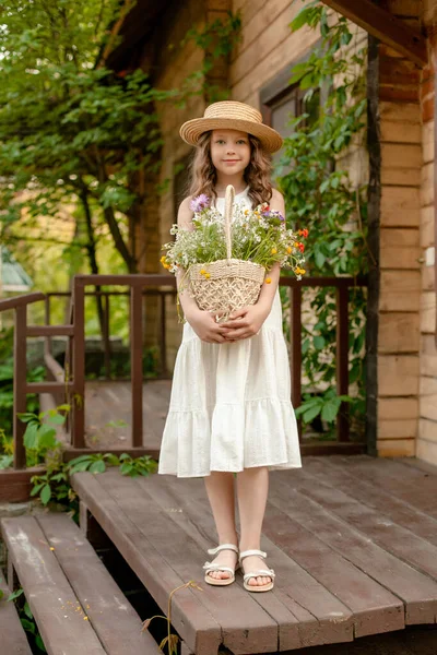 Modest preteen girl standing on country house threshold with basket of wildflowers