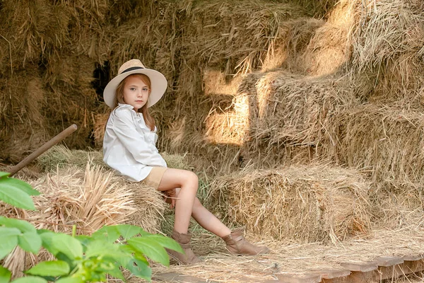 Маленькая девочка в деревенской одежде сидит на стоге сена на сеновале — стоковое фото