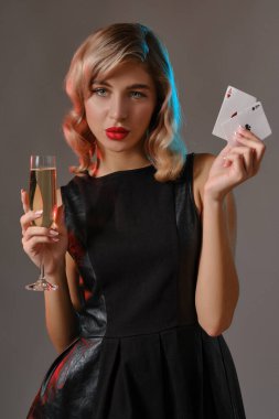 Siyah elbiseli sarışın kız elinde iki iskambil kâğıdı ve bir bardak şampanya ile gri arka planda poz veriyor. Kumar, poker, kumarhane. Yakın plan..