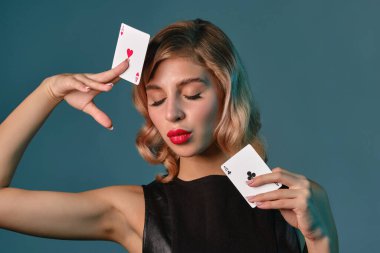 Siyah deri elbiseli sarışın kız mavi arka planda poz veren iki iskambil kağıdı gösteriyor. Kumar eğlencesi, poker, kumarhane. Yakın plan..