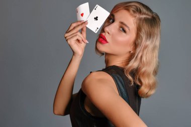 Siyah elbiseli sarışın kız iki iskambil kağıdı gösteriyor, gri arka planda yan poz veriyor. Kumar eğlencesi, poker, kumarhane. Yakın plan..