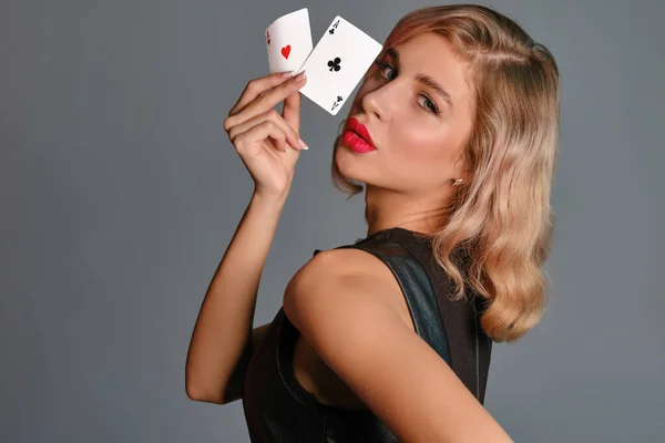 Chica rubia en vestido negro mostrando dos cartas de juego, posando de lado sobre fondo gris. Entretenimiento, póquer, casino. Primer plano. —  Fotos de Stock