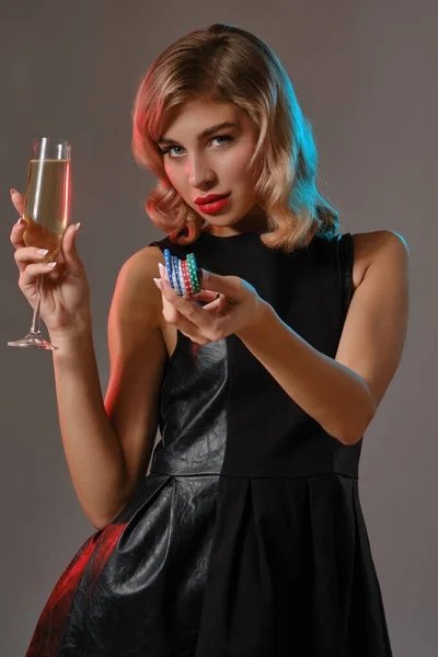 Blonde girl in black dress holding glass of champagne and chips, posing against gray background. Gambling, poker, casino. Close-up. Stock Photo