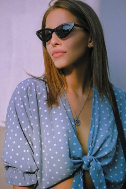 Uzun mavi elbiseli, güneş gözlüklü ve omzunda küçük siyah bir el çantası olan sarışın kız şehirde tek başına yürüyor. Moda ve tarz. Yakın çekim..