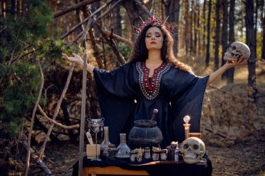 Siyah saçlı, uzun elbiseli, uzun kıvırcık saçlı bir cadı. Çam ormanında poz vermek. Kafatası tutuyorum. Büyü, büyü ve büyücülük. Yakın plan..