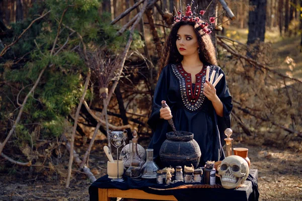 Belle sorcière en robe longue noire, avec couronne rouge dans ses longs cheveux bouclés. Poser dans la forêt de pins. Sortilèges, magie et sorcellerie. Gros plan. — Photo