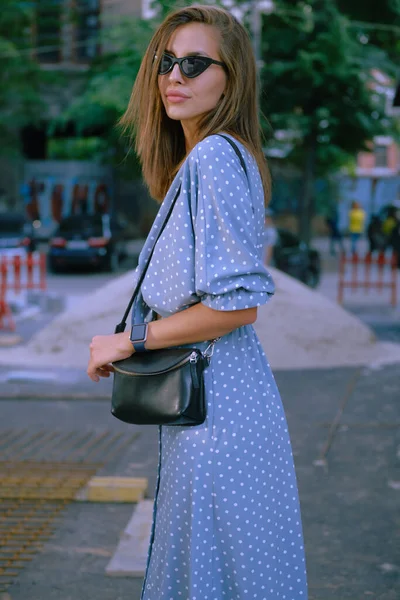 Menina loira em vestido azul longo, óculos de sol e uma pequena bolsa preta em seu ombro está andando sozinha na cidade. Moda e estilo. Imagem de close-up. — Fotografia de Stock