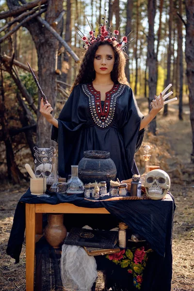 Belle sorcière en robe longue noire, avec couronne rouge dans ses longs cheveux bouclés. Poser dans la forêt de pins. Sortilèges, magie et sorcellerie. Gros plan. — Photo