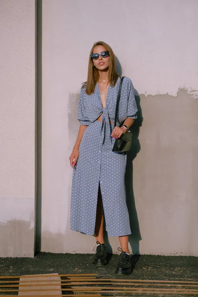 Menina loira em vestido azul longo, óculos de sol e uma pequena bolsa preta em seu ombro está andando sozinha na cidade. Moda e estilo. Comprimento total. — Fotografia de Stock