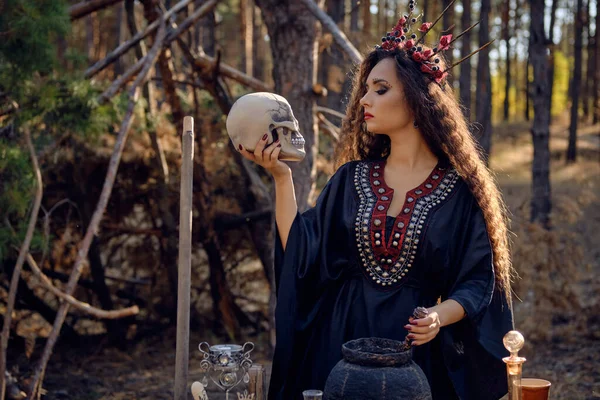 Sorcière en robe longue noire, avec couronne rouge dans ses longs cheveux bouclés. Poser dans la forêt de pins. Tenir le crâne. Sortilèges, magie et sorcellerie. Gros plan. — Photo