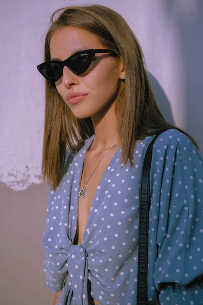 Menina loira em vestido azul longo, óculos de sol e uma pequena bolsa preta em seu ombro está andando sozinha na cidade. Moda e estilo. Imagem de close-up. — Fotografia de Stock