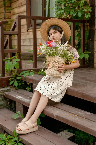 Donkerharig meisje zittend op de stoep van landelijk huis met mand van wilde bloemen — Stockfoto