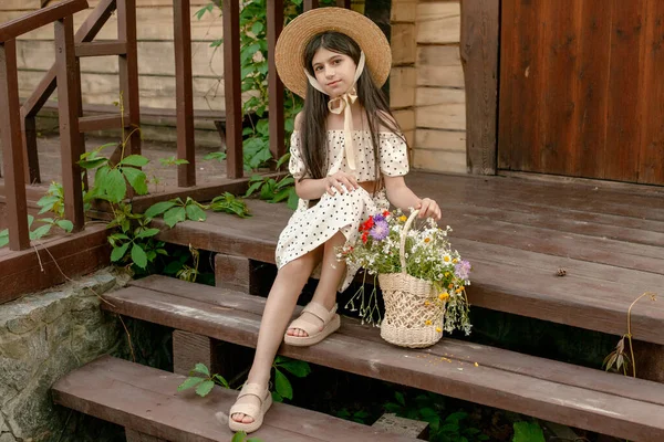 Темноволосая девочка-подросток сидит на деревянном пороге с корзиной полевых цветов — стоковое фото